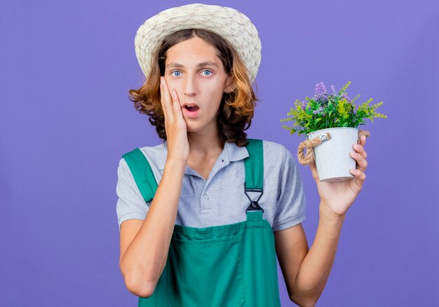 びっくりしている鉢植えの植物を保持しているジャンプスーツと帽子を身に着けている若い庭師の男