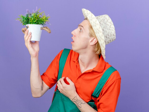 Young gardener man wearing jumpsuit and hat holding potted plant looking at it surprised