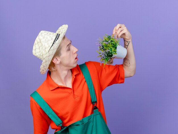 Young gardener man wearing jumpsuit and hat holding potted plant looking at it surprised