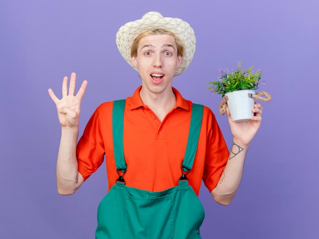 笑顔の鉢植えの植物を保持しているジャンプスーツと帽子を身に着けている若い庭師の男