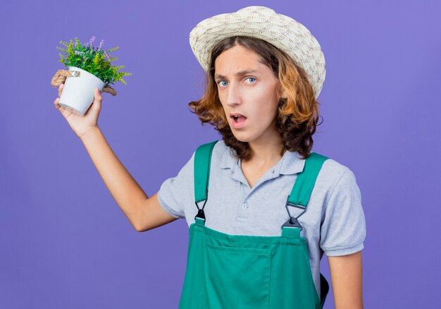 Молодой садовник в комбинезоне и шляпе держит горшечное растение с сердитым