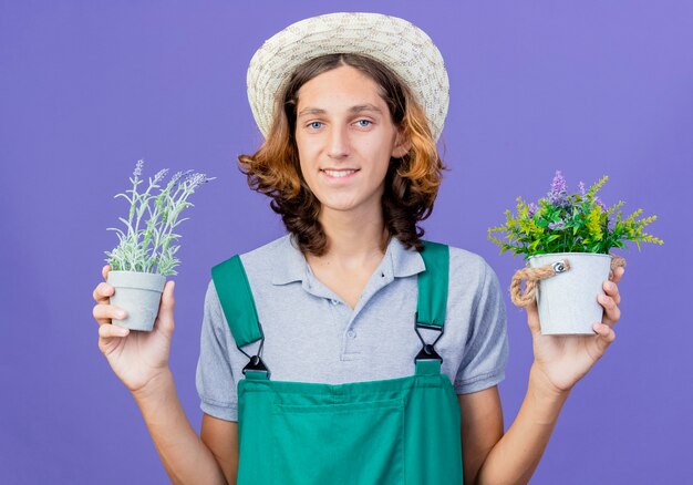 笑顔の鉢植えの植物を保持しているジャンプスーツと帽子を身に着けている若い庭師の男