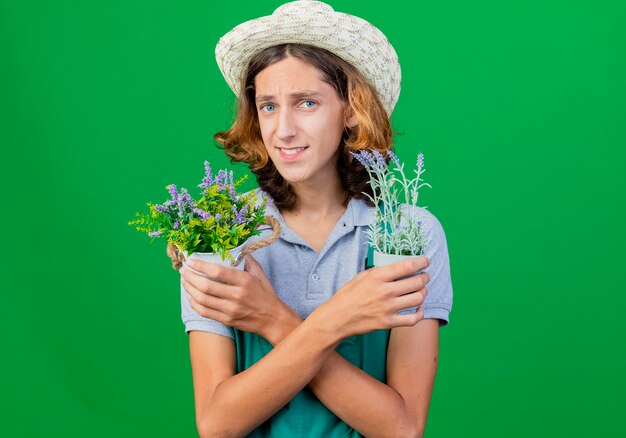 Free photo young gardener man wearing jumpsuit and hat holding potted plants smiling