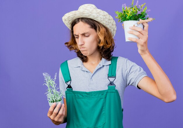 Young gardener man wearing jumpsuit and hat holding potted plants