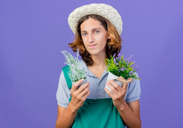 Young gardener man wearing jumpsuit and hat holding potted plants