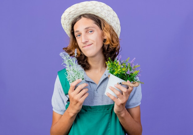 Young gardener man wearing jumpsuit and hat holding potted plants