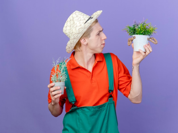 Young gardener man wearing jumpsuit and hat holding potted plants