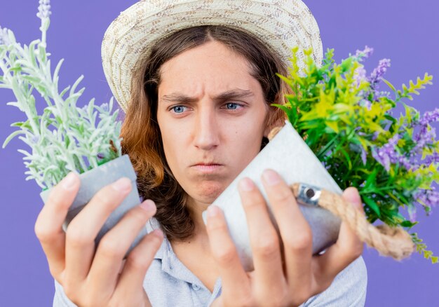 Young gardener man wearing jumpsuit and hat holding potted plants