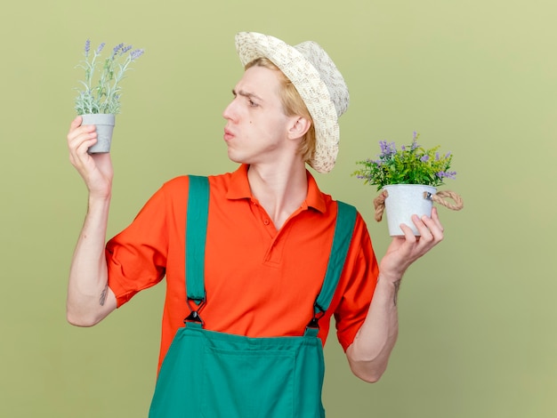 Young gardener man wearing jumpsuit and hat holding potted plants