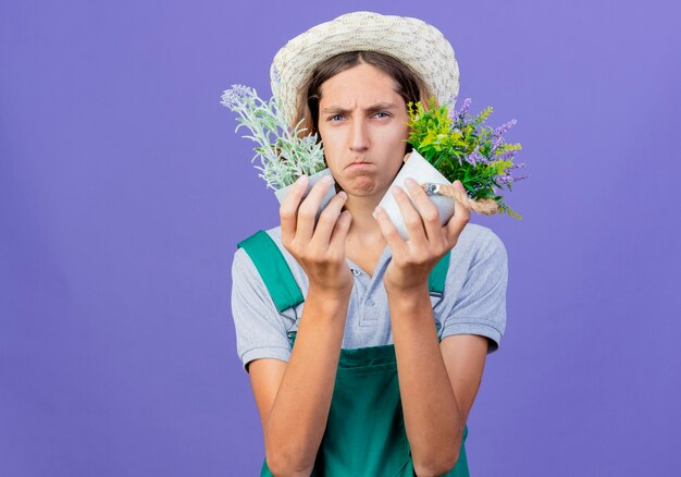 Young gardener man wearing jumpsuit and hat holding potted plants