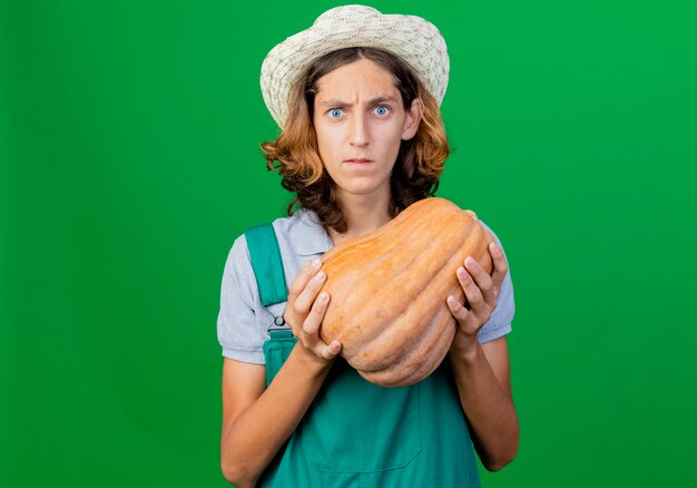 Young gardener man wearing jumpsuit and hat holding pumpkin being confused