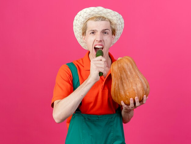 Young gardener man wearing jumpsuit and hat holding pumpkin and cucumber