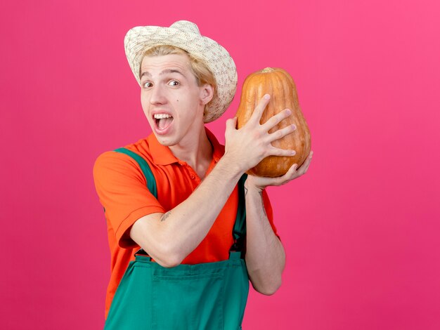 Young gardener man wearing jumpsuit and hat holding pumpkin happy and excited