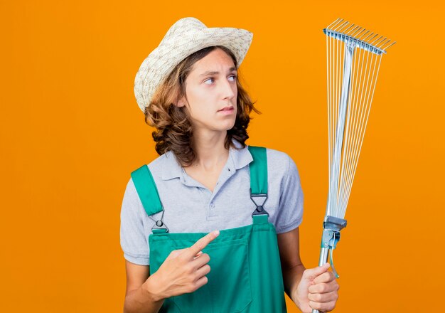 Young gardener man wearing jumpsuit and hat holding rake pointing with index finger