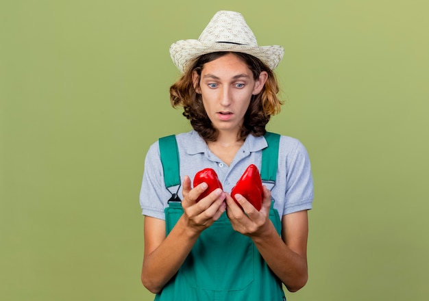 Free photo young gardener man wearing jumpsuit and hat holding red bell peppers