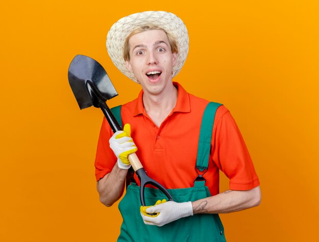 Young gardener man wearing jumpsuit and hat holding shovel being surprised