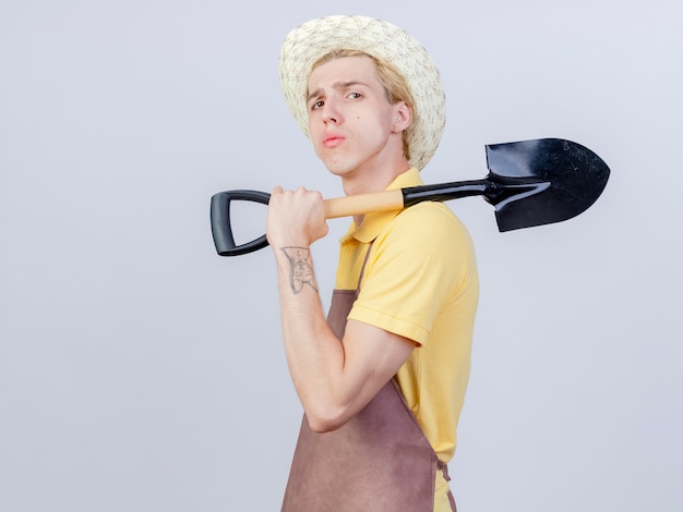 Young gardener man wearing jumpsuit and hat holding shovel on his shoulder looking confident