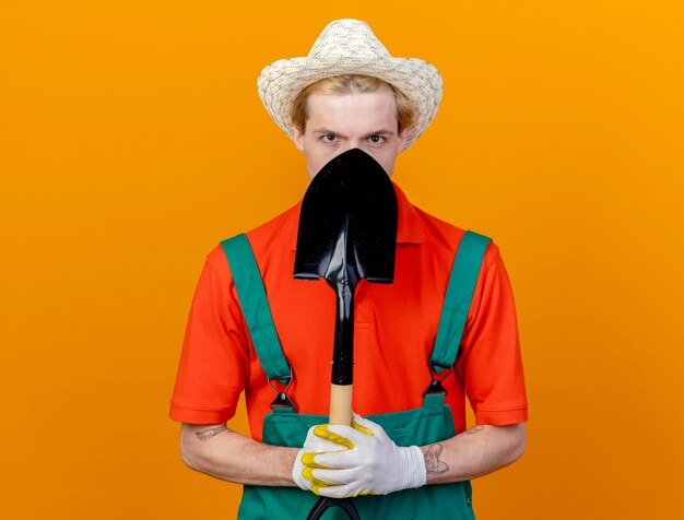 Young gardener man wearing jumpsuit and hat holding shovel looking at camera hiding his face standing over orange background