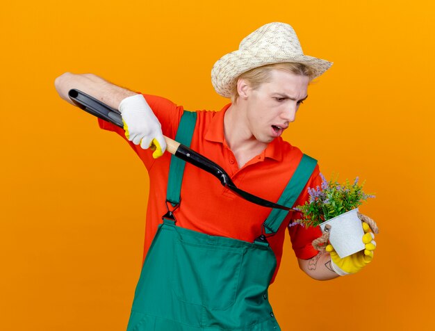 Young gardener man wearing jumpsuit and hat holding shovel and potted plant