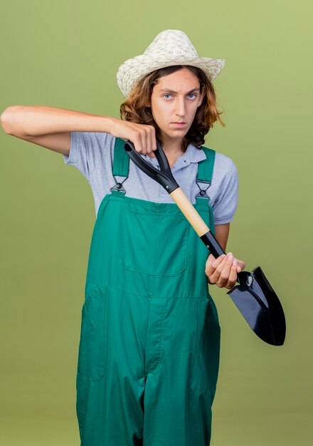 Young gardener man wearing jumpsuit and hat holding shovel