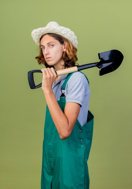 Young gardener man wearing jumpsuit and hat holding shovel