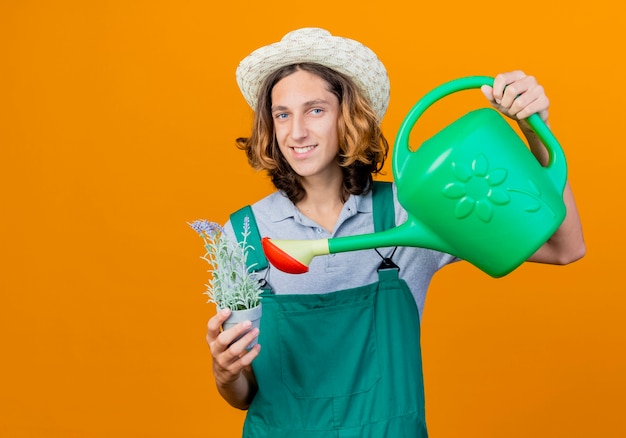 Young gardener man wearing jumpsuit and hat holding watering can and potted plant