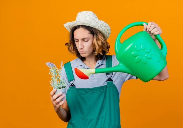 Young gardener man wearing jumpsuit and hat holding watering can and potted plant