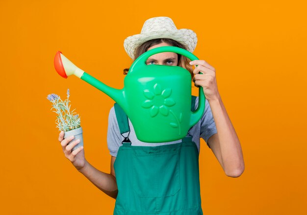 Free photo young gardener man wearing jumpsuit and hat holding watering can and potted plant