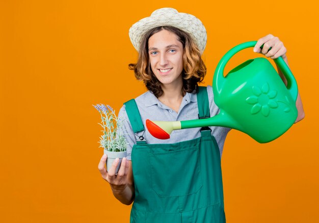 Young gardener man wearing jumpsuit and hat holding watering can