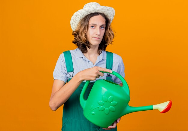 Young gardener man wearing jumpsuit and hat holding watering can