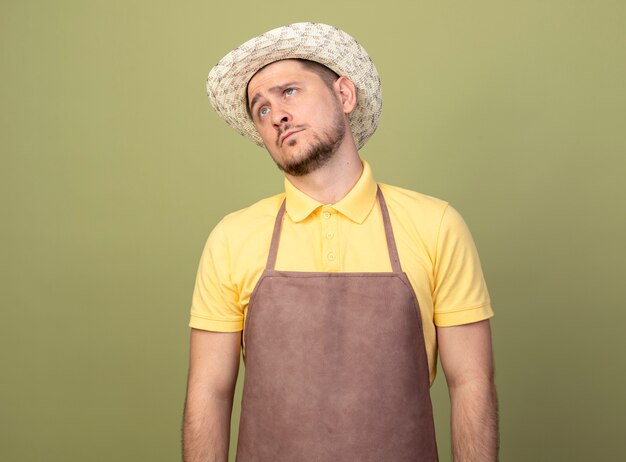 Giovane giardiniere uomo che indossa tuta e cappello guardando da parte perplesso con l'espressione triste sul viso in piedi sopra la parete chiara