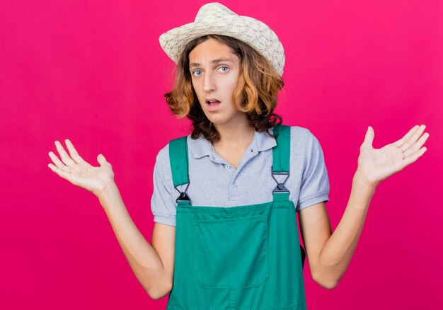 Young gardener man wearing jumpsuit and hat looking confused