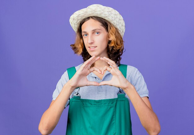 Young gardener man wearing jumpsuit and hat making heart gesture with fingers smiling