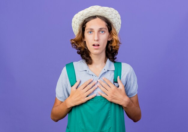 Young gardener man wearing jumpsuit and hat pointing at himself