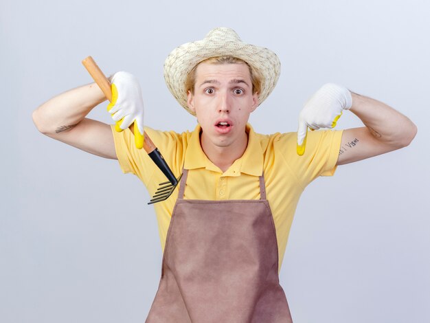 Young gardener man wearing jumpsuit and hat in rubber gloves holding mini rake being surprised pointing down 