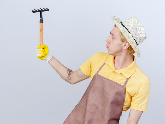 Young gardener man wearing jumpsuit and hat in rubber gloves holding mini rake looking at it with serious face 