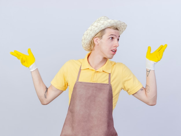Young gardener man wearing jumpsuit and hat in rubber gloves looking aside spreading hands to the sides smiling 