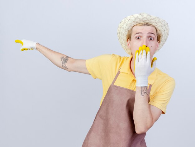 Young gardener man wearing jumpsuit and hat in rubber gloves pointing with index finger at something being shocked 