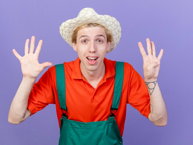 Young gardener man wearing jumpsuit and hat showing number nine