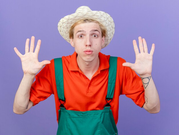 young gardener man wearing jumpsuit and hat showing number ten being surprised