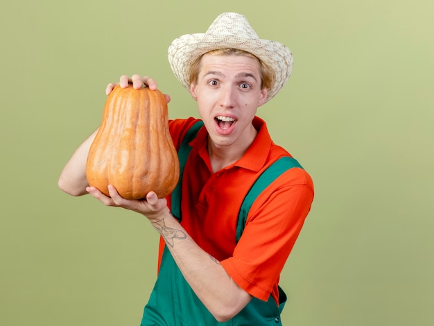 Free photo young gardener man wearing jumpsuit and hat showing pumpkin