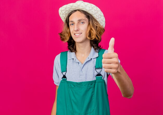 Young gardener man wearing jumpsuit and hat smiling showing thumbs up