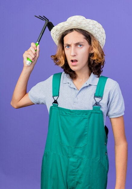 Young gardener man wearing jumpsuit and hat swinging a mattock