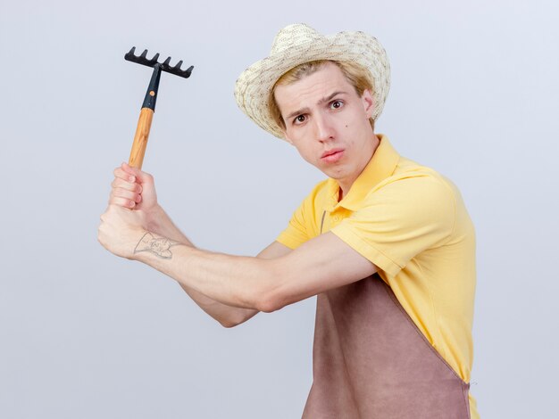 Young gardener man wearing jumpsuit and hat swinging mini rake being displeased 