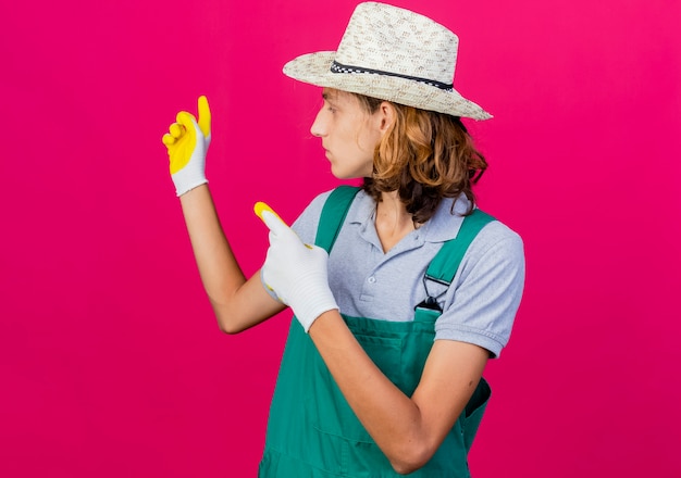 Young gardener man wearing jumpsuit and hat wearing rubber gloves pointing