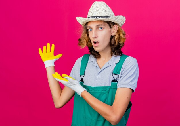 Young gardener man wearing jumpsuit and hat wearing rubber gloves presenting