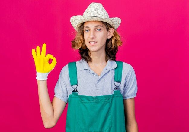 Young gardener man wearing jumpsuit and hat wearing rubber gloves smiling showing ok sign