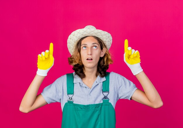 Young gardener man wearing jumpsuit and hat wearing rubber gloves