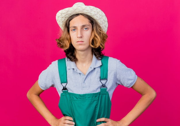 Free photo young gardener man wearing jumpsuit and hat with serious face with arms at hip