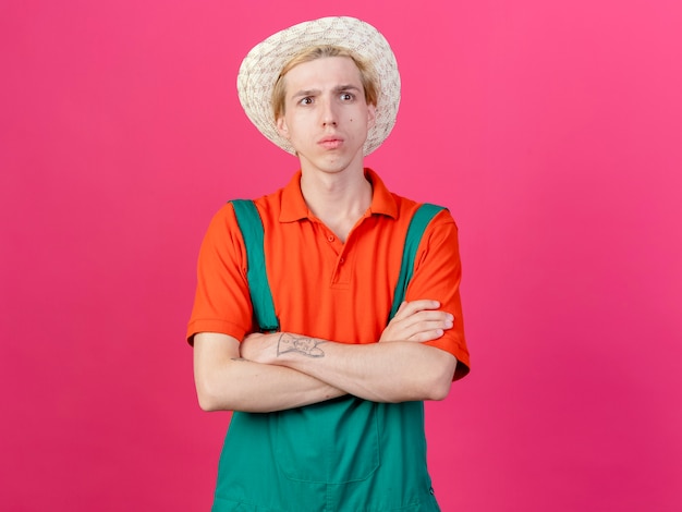 Young gardener man wearing jumpsuit and hat with serious face with crossed hands on chest